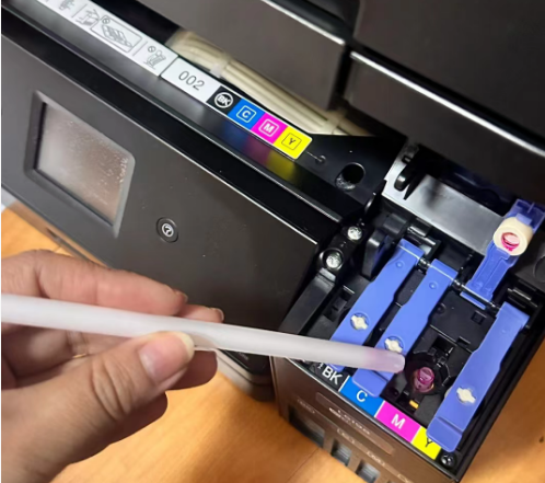 A close-up view of a person carefully using a syringe with a tube to apply cleaning solution to an Epson printhead nozzle.